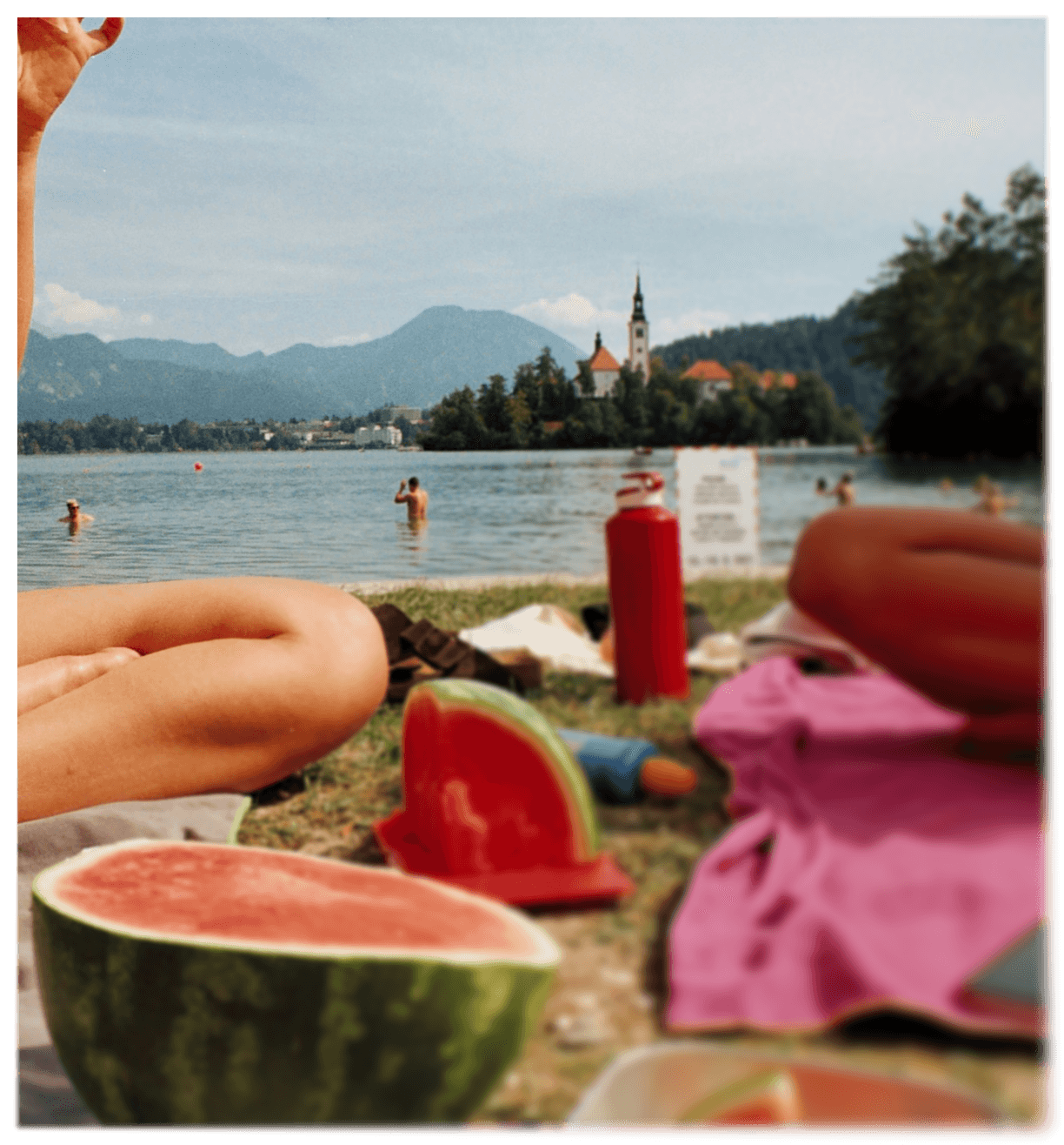 Lakeside picnic scene with watermelon, towels, and a mountain lake with an island church in the background